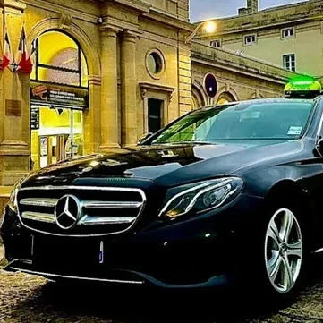 Taxi Drive It Saint-Quentin-sur-Isère – transfert ski Alpe d’Huez Taxi noir de marque Mercedes-Benz devant un bâtiment historique avec des drapeaux.