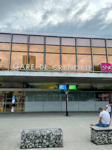 Gare de Grenoble avec des panneaux d'information et des voyageurs assis sur un banc.