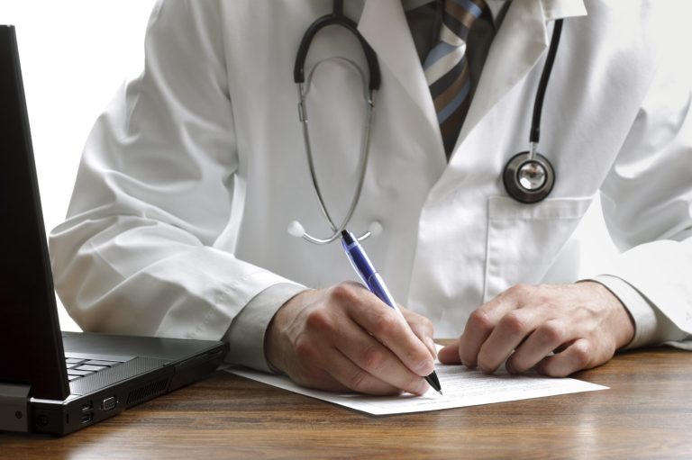 Médecin en blouse blanche prenant des notes près d'un ordinateur portable.